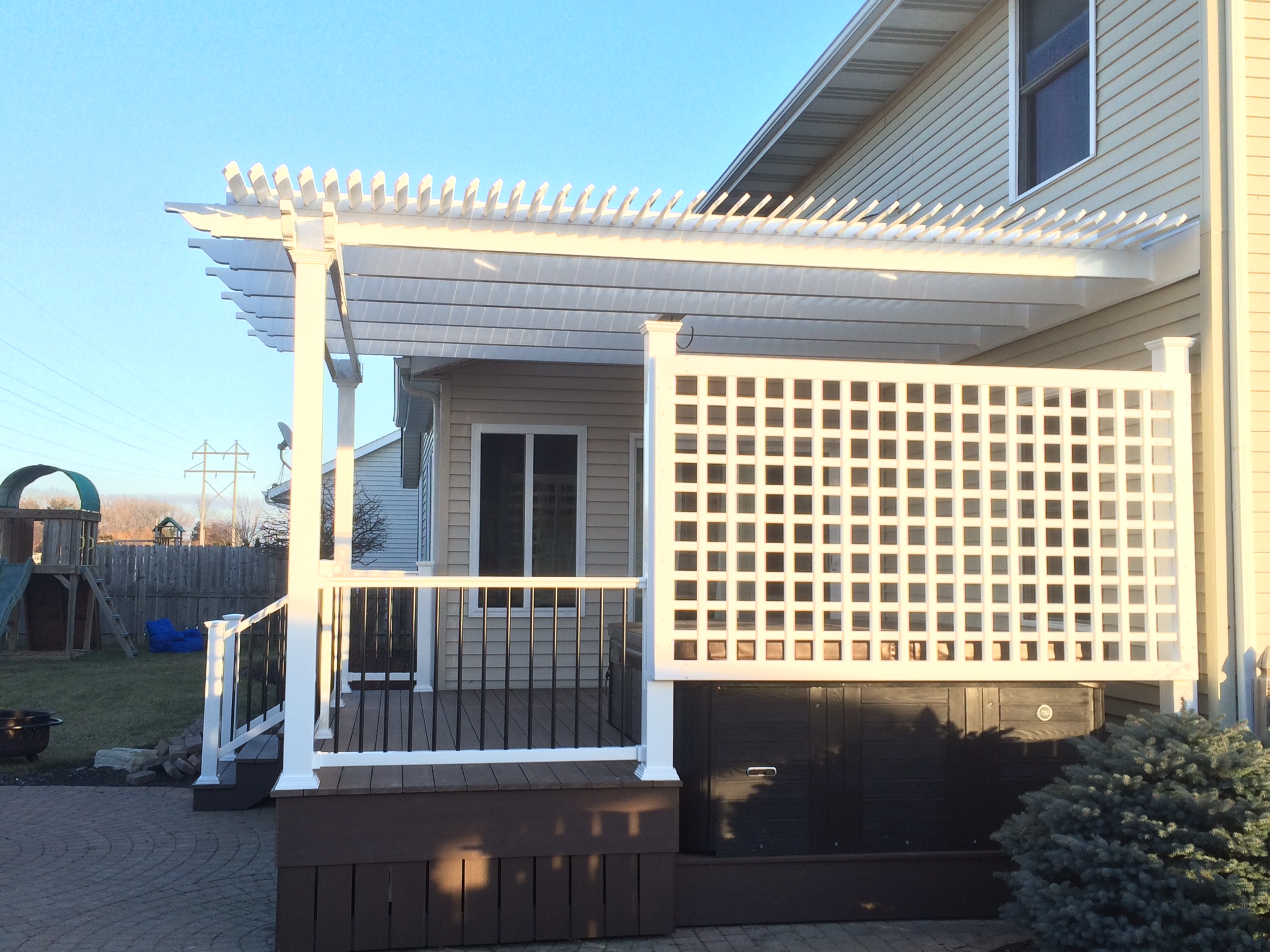 This two-story home with tan siding has a white pergola attached to the porch. There is a privacy trellis on the side of the deck, and there is a playset in the backyard.