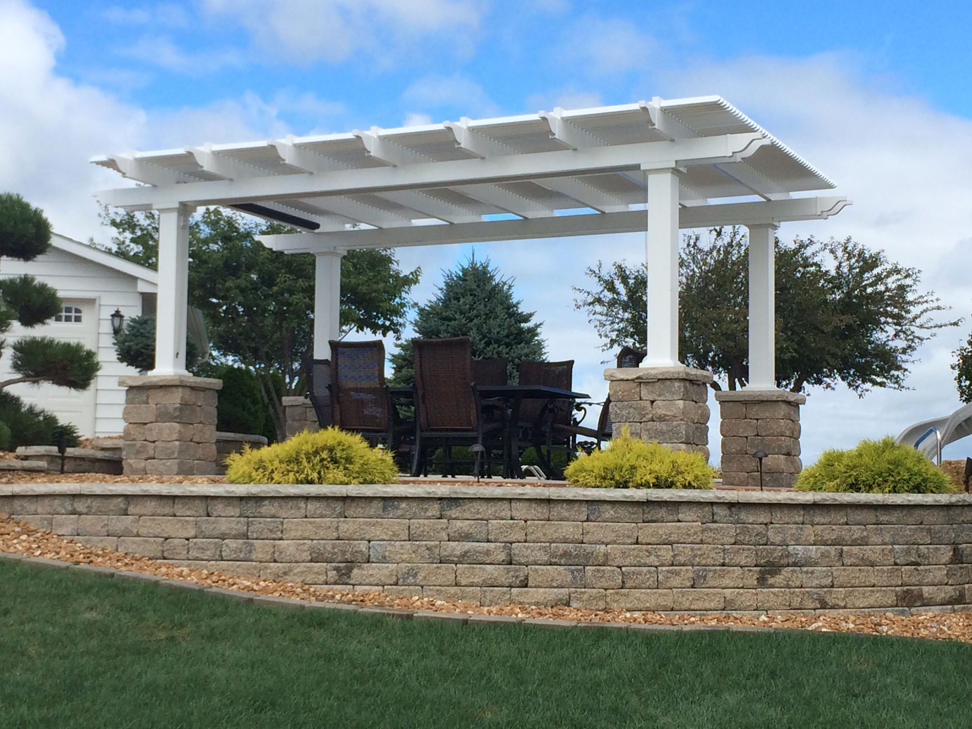This white pergola stands alone above an outdoor eating area. There are green shrubs on the sides of the poles, and there are trees in the yard.