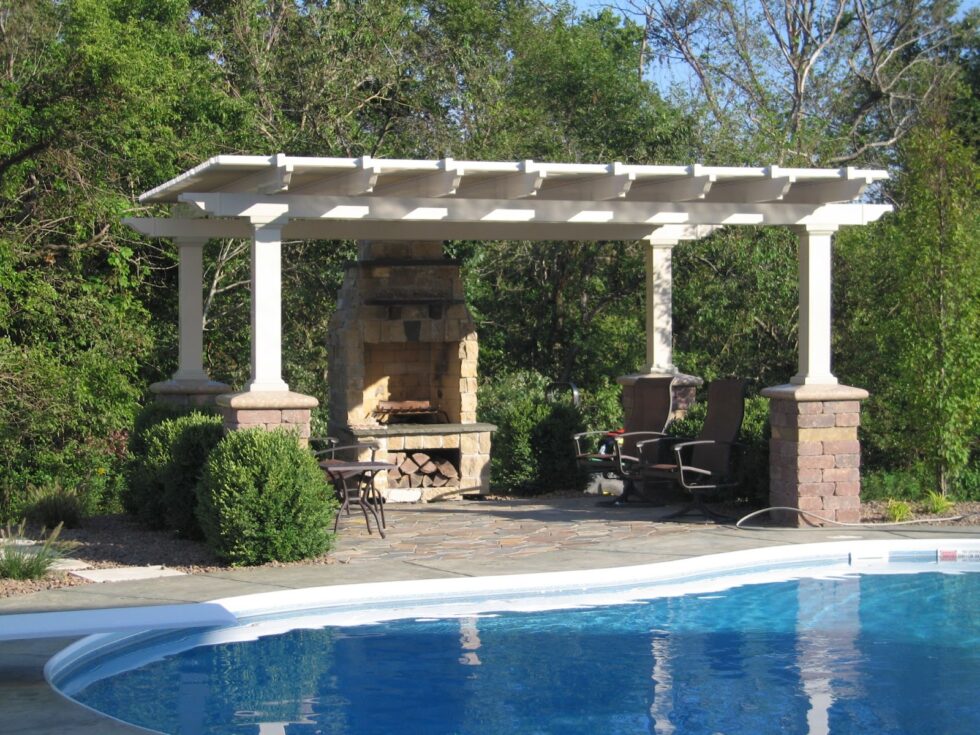 This outdoor space has a pool with blue water and white siding. There is a seating area with a white pergola and a stone fireplace. There are a few chairs under the pergola, and there are trees behind it.