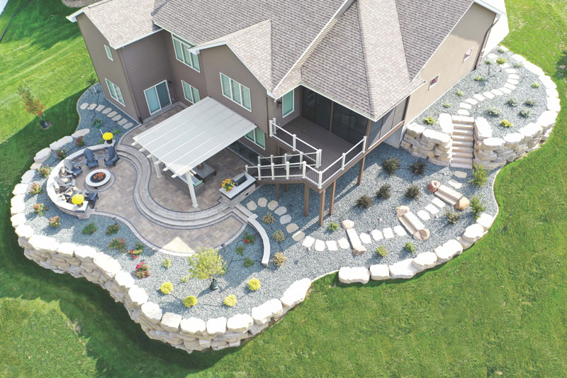Beautifully designed backyard aerial view, with stone retaining wall, stone patio, elveated deck and a shade pergola.