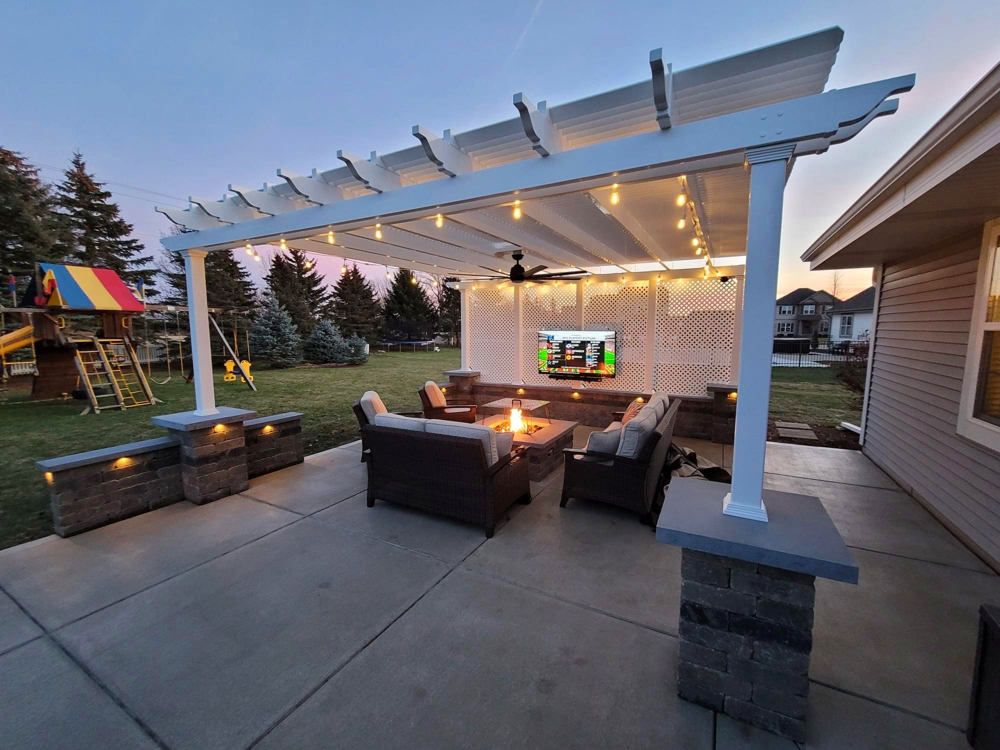 Outdoor living patio with tv, firepit and furniture under a large white pergola on stone columns