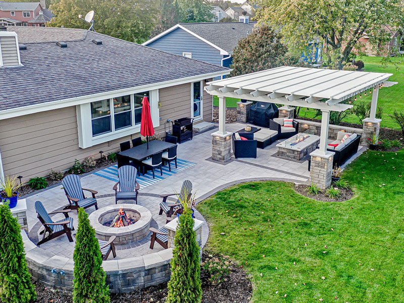 Stone patio in a backyard with a pergola on stone columns over an outdoor seating living area and fire table.