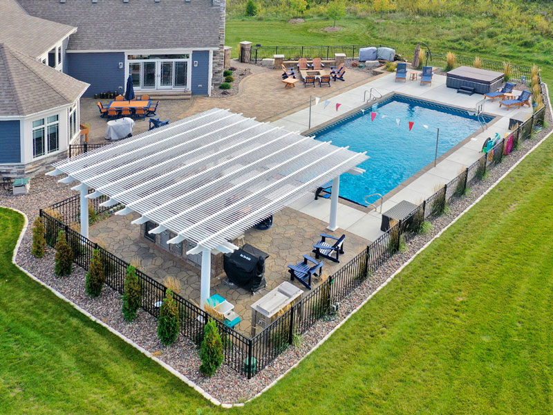 Aerial view of a large white pergola over a outdoor kitchen and bar next to a large inground swimming pool and lovely landscaped yard.