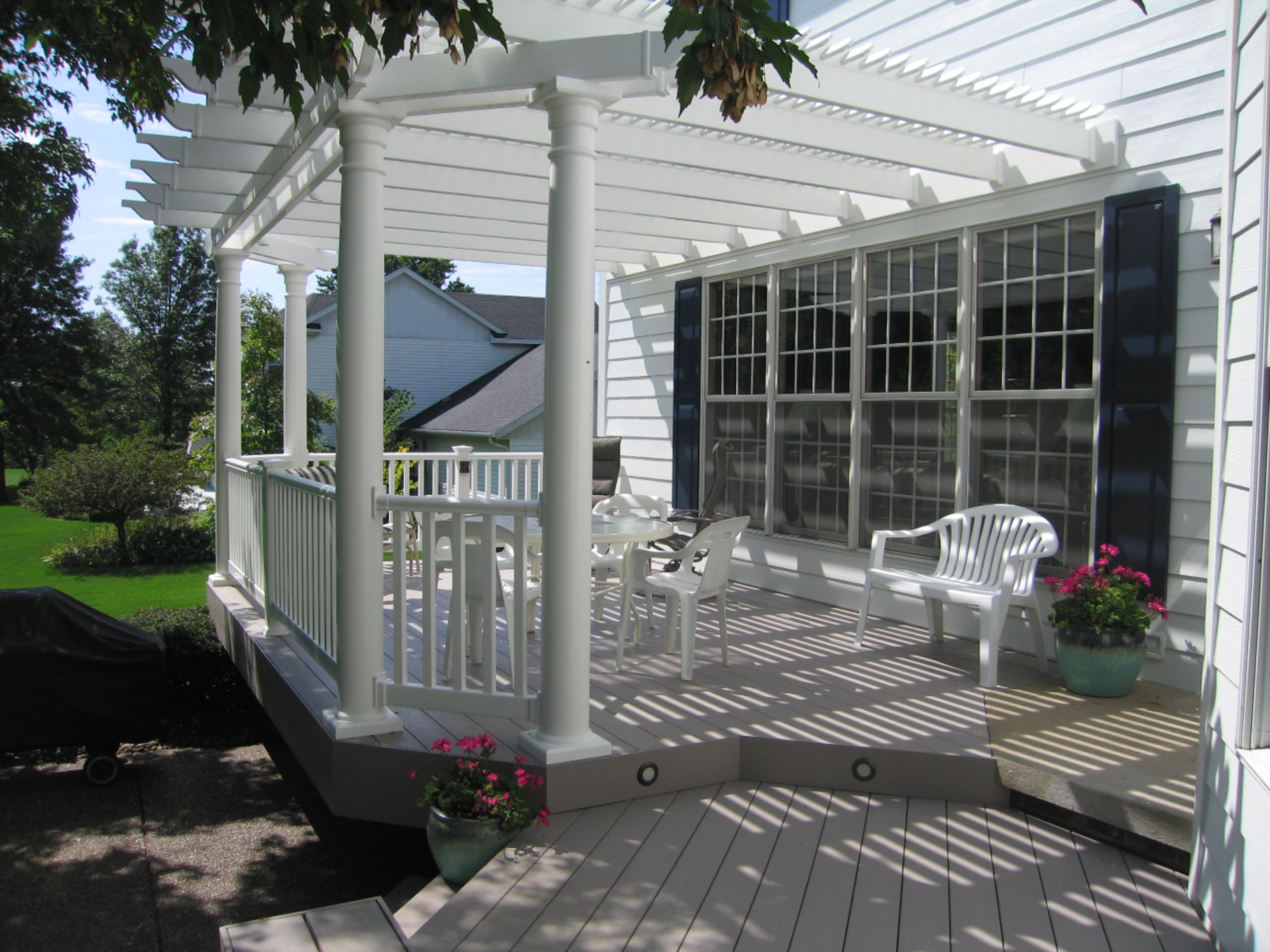 This white house has four large windows looking out to the deck area. There is a white pergola connected to the deck and the house, covering white deck furniture and pink flowers in blue flower pots.
