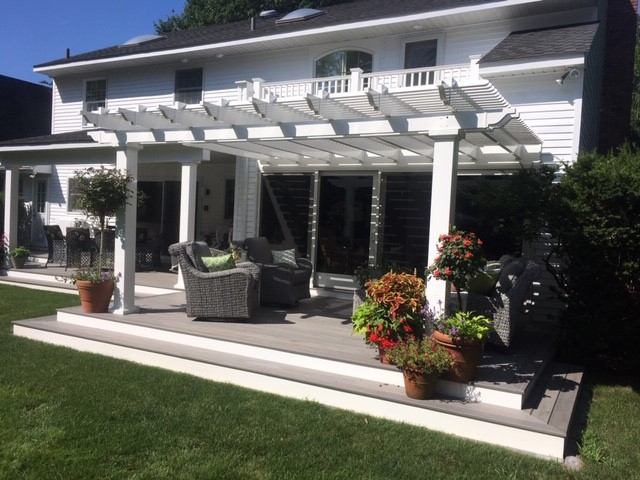 A white home installed a pergola to cover their outdoor patio space. There are two grey chairs with green pillows under the pergola, and there are potted plants next to the pergola.