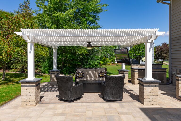 Side view of a white vinyl freestanding pergola on a paved patio. The pergola has a fan and tv attached and sits above outdoor seating area.