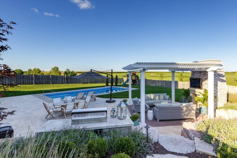 Nicely landscaped back patio with freestanding pergola, tv and outdoor fireplace next to a pool