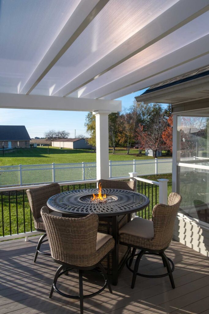 A white vinyl pergola with white polycarbonate roof over a deck with a fire table and chairs.