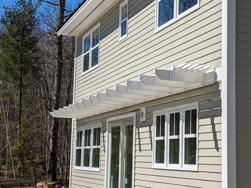 New construction home with a white vinyl eyebrow pergola running the length of the back of a house over windows and patio doors.