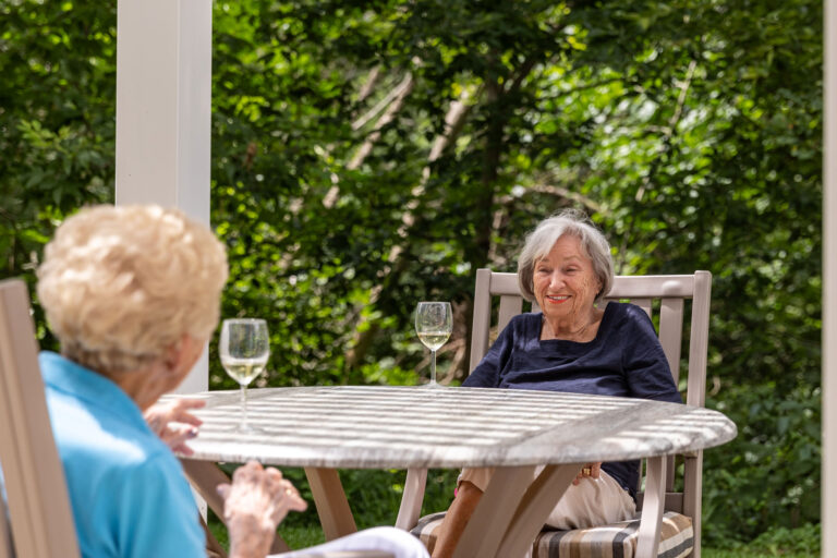 two older women sit talking and sipping wine while sitting outdoors on the patio of their senior living facility