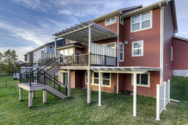 Two pergolas, one over a raised deck and a second one attached to the home above grass area on the back of a red house.